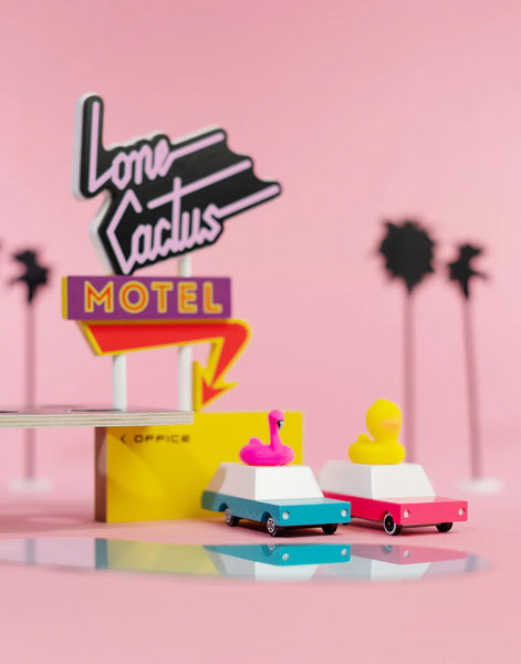 Model cars with flamingo and duck decorations in front of a 'Lone Cactus Motel' sign on a pink background.