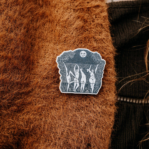 Decorative brooch with a design of people dancing in front of a moon on a brown textured cardigan.