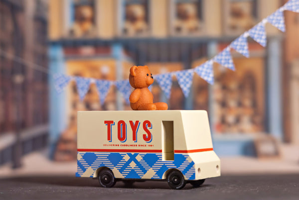 Toy truck with a teddy bear on top, labeled 'TOYS', in front of a blurred store facade.