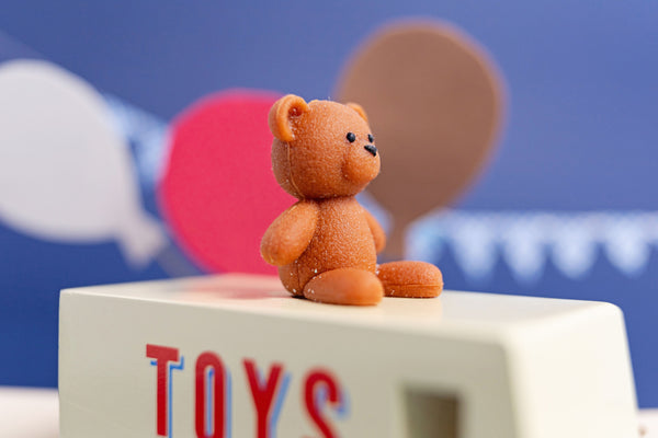 Small brown teddy bear on a block with 'TOYS' letters against a blue background