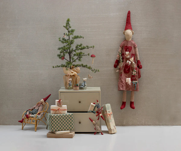 Decorative scene with small tree, gift boxes, and pixy figurines against a plain wall.