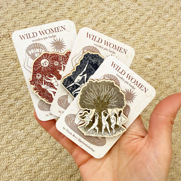 Three 'Wild Women' wooden pin badges on cards held by a hand against a beige carpet background.