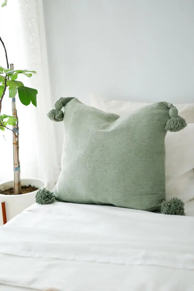 Green pillow with tassels on a bed, with a plant in the background.