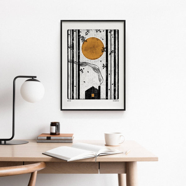 Framed artwork on a wall above a desk with a lamp and books.