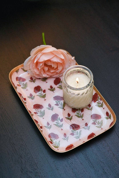 A small pink floral tray with a lit candle in a clear glass jar and a pale pink flower, arranged on a dark wooden surface.