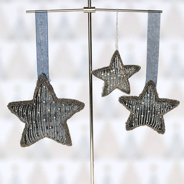 Three beaded star-shaped ornaments in a metallic silver tone, each hanging from a light blue ribbon on a metal display stand against a softly blurred white background.