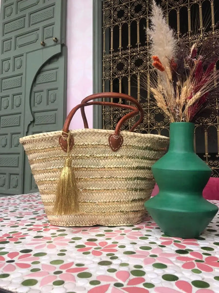 Woven basket with brown handle and tassel next to a green vase on a floral tablecloth.