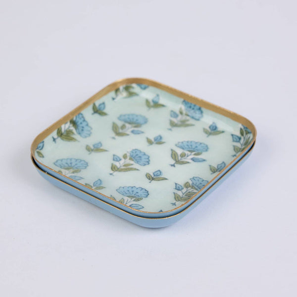 Two enamelled iron coasters with rounded corners, featuring a light blue and white floral pattern with gold trim, set against a plain white background.
