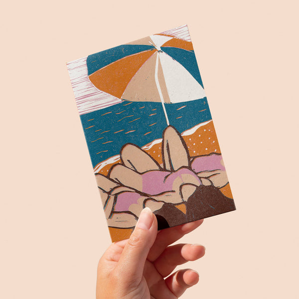 Hand holding a small rectangular card with a colourful design of an umbrella and women on a beach on a beige background