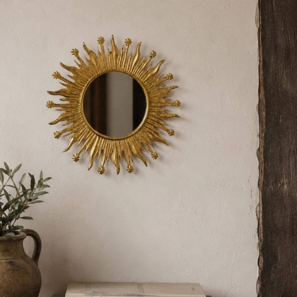 Decorative gold sunburst mirror on a wall with a plant on a surface in the foreground.