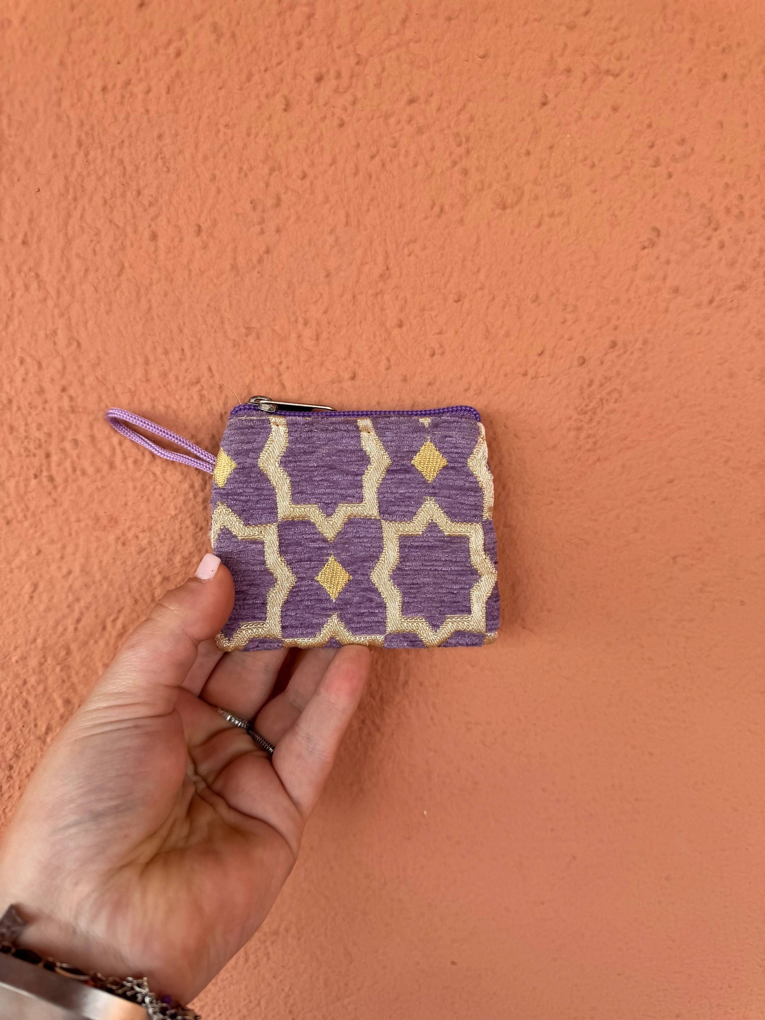 Hand holding a small purple and beige patterned pouch against an orange wall.