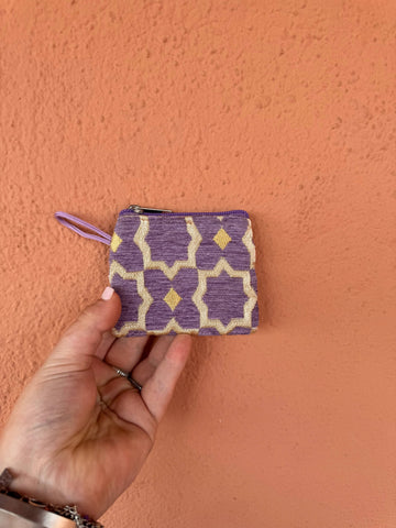 Hand holding a small purple and beige patterned pouch against an orange wall.