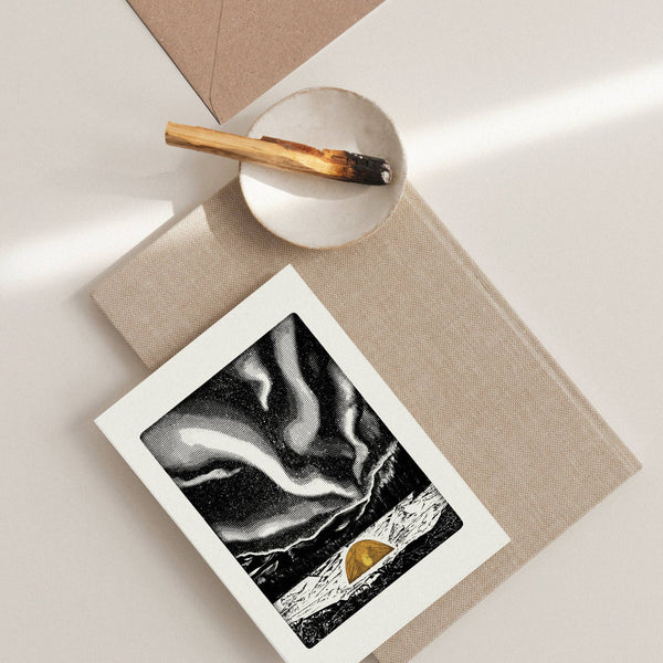 A greeting card with a white border showing a black-and-white abstract landscape and a small orange tent, styled on a beige surface with a matching envelope, and a small dish.