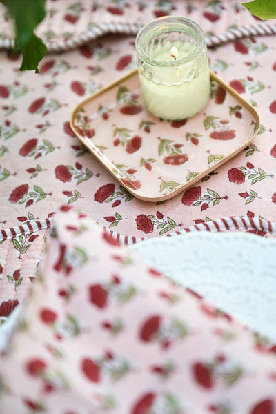 A pink floral blanket with red roses and green leaves, styled with a gold rimmed square coaster/tray holding a small lit glass jar candle.