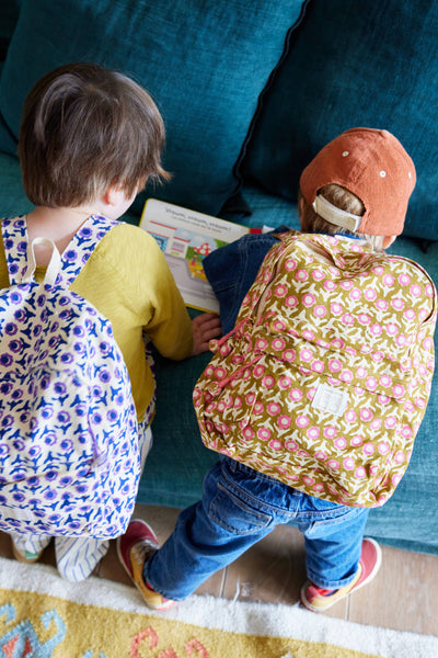 Two children with backpacks leaning on a blue couch reading a book.
