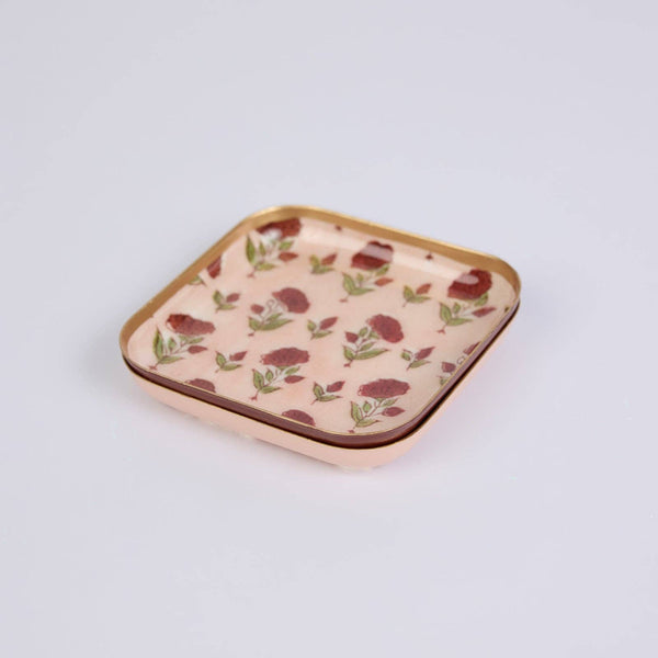 A small square coaster/jewellery tray with rounded corners, featuring a light pink floral pattern with red rose motifs and green leaves, framed by a metallic gold border.