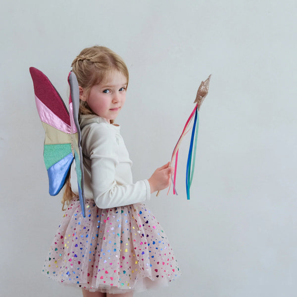Child wearing colorful fairy wings and holding a wand against a plain background