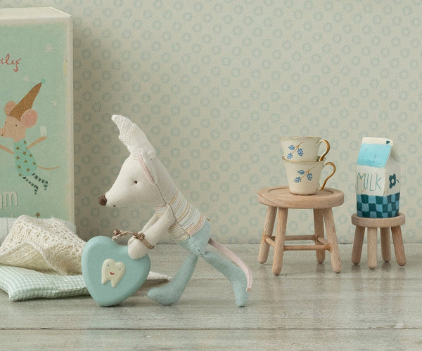 Stuffed toy mouse with a heart-shaped tooth tin in a room with a patterned wall and small wooden stool with cups and milk carton.