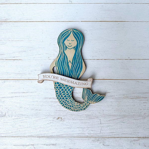 A wooden mermaid-shaped magnet with teal and cream painted details, featuring a banner that reads 'YOU'RE MERMAZING,' displayed on a white-washed wood plank background.