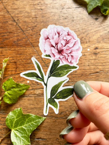 Hand holding a pink flower sticker on a wooden surface with green leaves.