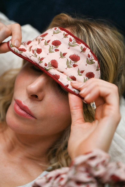 A woman wearing a light pink floral sleep mask, holding it in place with both hands in a close-up lifestyle shot.