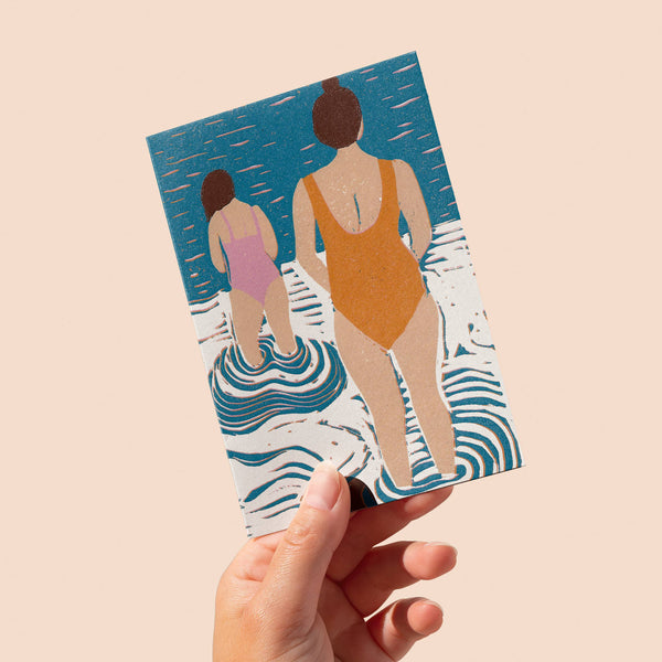 Card with illustration of two people in swimsuits on a wavy background, held by a hand against a beige background.