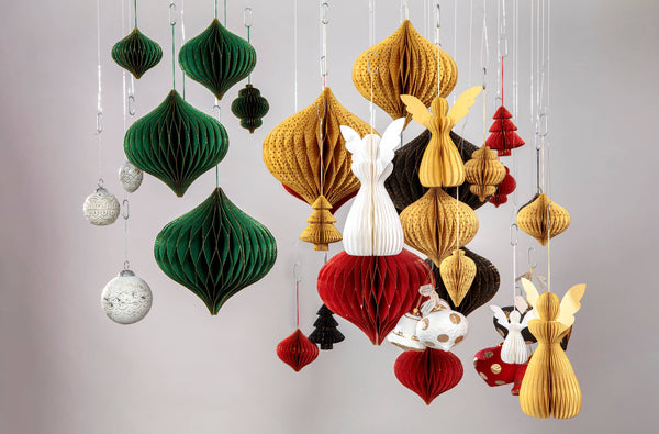 A collection of hanging paper honeycomb ornaments in green, gold, red, white, and black, including teardrop shapes, small Christmas tree ornaments, and angel figures, suspended against a neutral grey background.
