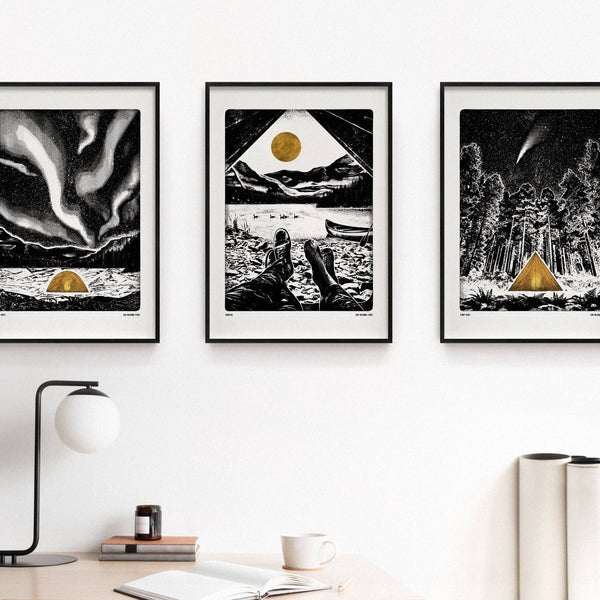 Three framed artworks with a camping theme on a wall with a desk and lamp in the foreground.