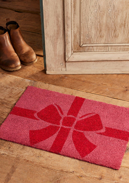 A pink coir doormat with a red gift box and bow design, placed on a wooden floor next to a pair of brown ankle boots and a rustic wooden door.