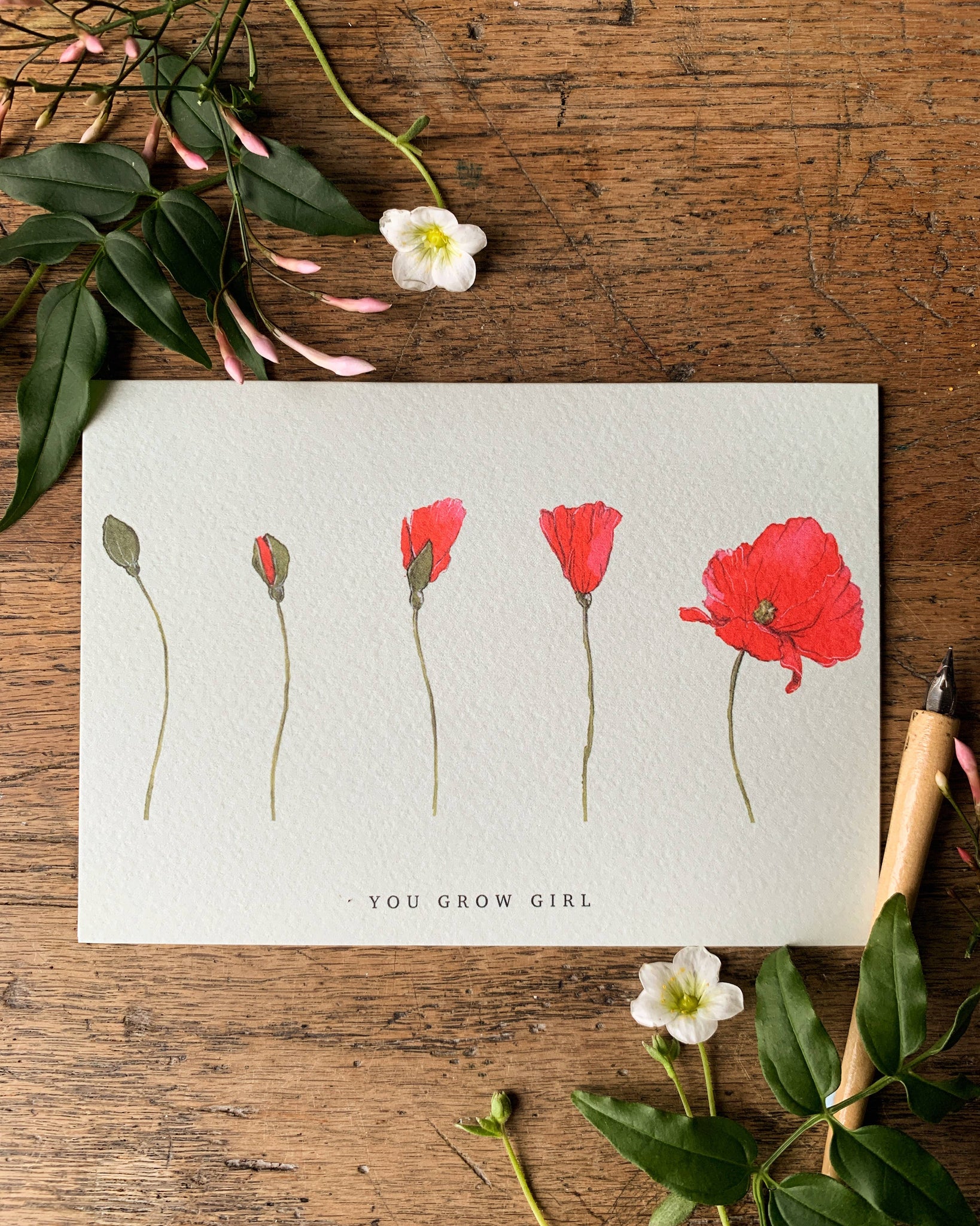 Card with red flowers and 'You Grow Girl' text on a wooden surface with flowers and leaves.