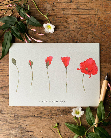 Card with red flowers and 'You Grow Girl' text on a wooden surface with flowers and leaves.
