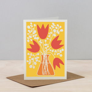 Greeting card with red tulips and white flowers on a yellow background, placed on a wooden surface with a gray background.