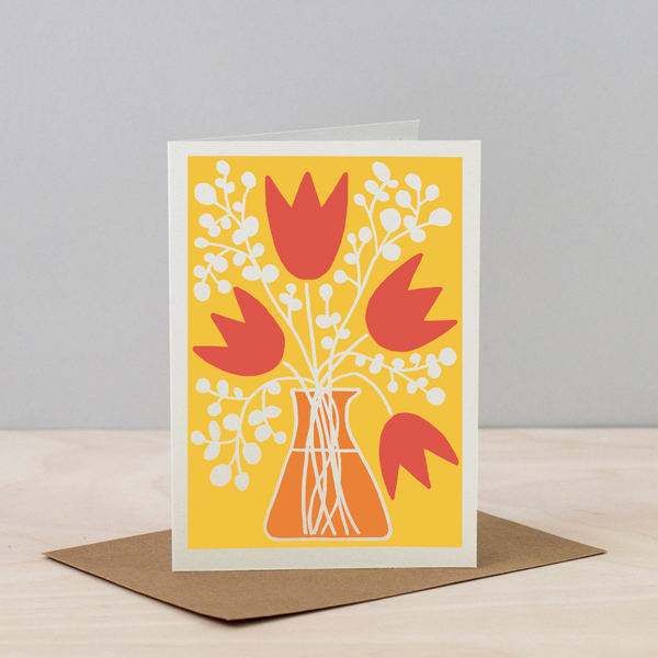 Greeting card with red tulips and white flowers on a yellow background, placed on a wooden surface with a gray background.