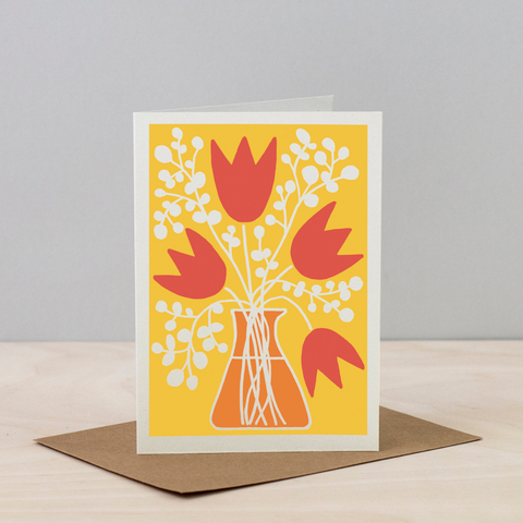 Greeting card with red tulips and white flowers on a yellow background, placed on a wooden surface with a gray background.