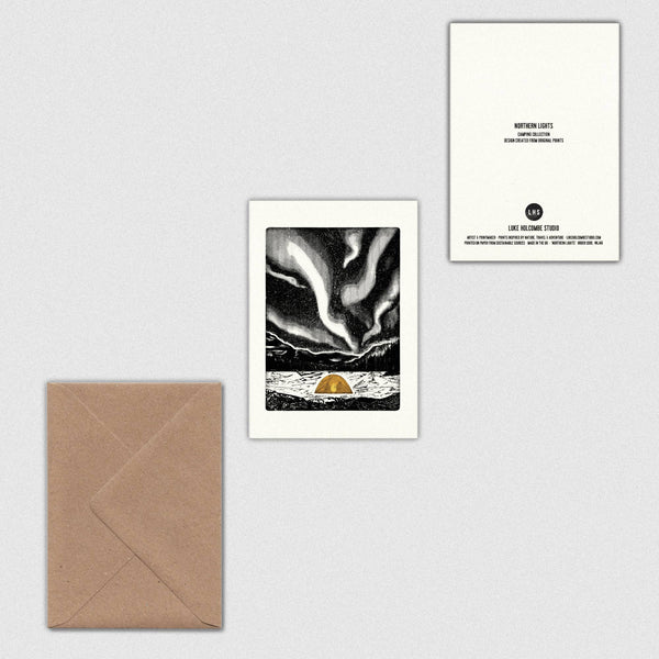 A minimalist art print card featuring a black and white northern lights illustration with a small orange tent at the bottom, shown alongside a brown kraft envelope and the back of the card on a light grey background.