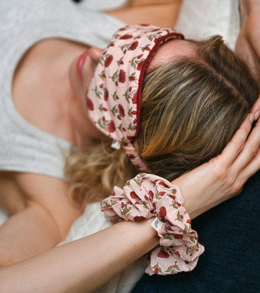 A close-up lifestyle shot of a woman resting her head on a pillow, wearing a matching pink floral sleep mask and scrunchie set.
