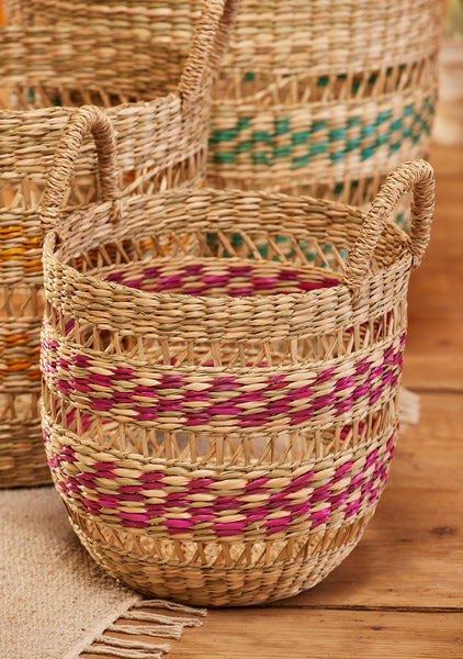 A woven natural fiber basket with two loop handles, decorated with horizontal bands of magenta and beige, displayed on a rustic wooden surface with other baskets in the background.