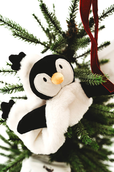 A plush penguin soother wearing a white fluffy hat and scarf, hanging on a green Christmas tree with a red ribbon.