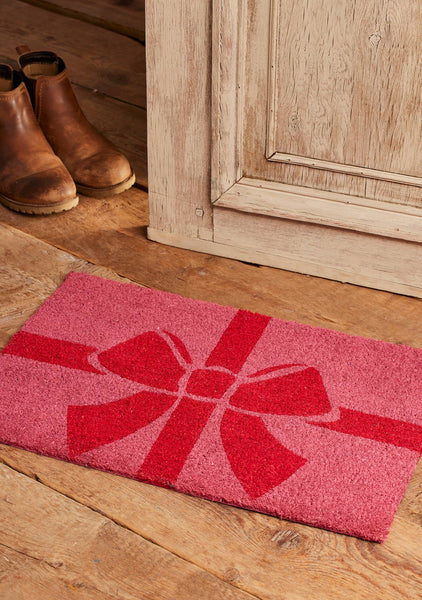 A pink coir doormat with a red gift box and bow design, placed on a rustic wooden floor next to a light wood cabinet and a pair of brown boots.
