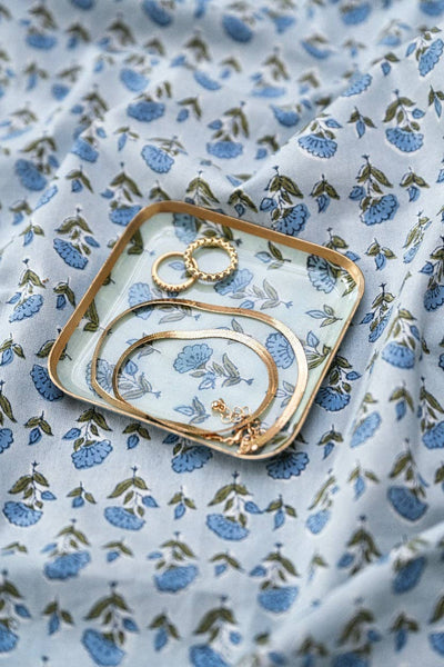 Gold-toned jewellery pieces, including rings and bracelets, arranged on a small square enamelled iron coaster/tray with a blue and white floral print, set on matching patterned fabric.