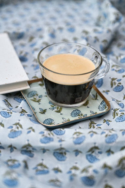 A clear glass cup filled with black coffee and a light tan crema sits on a square coaster with a blue floral pattern, placed on a matching blue floral fabric surface next to a white book.