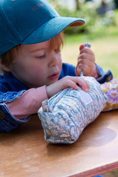 Child wearing a blue cap and jacket, interacting with a fabric pencil case on a wooden surface outdoors.