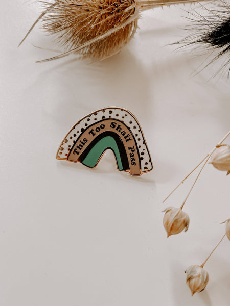 Rainbow-shaped pin with text on a light background with dried plants