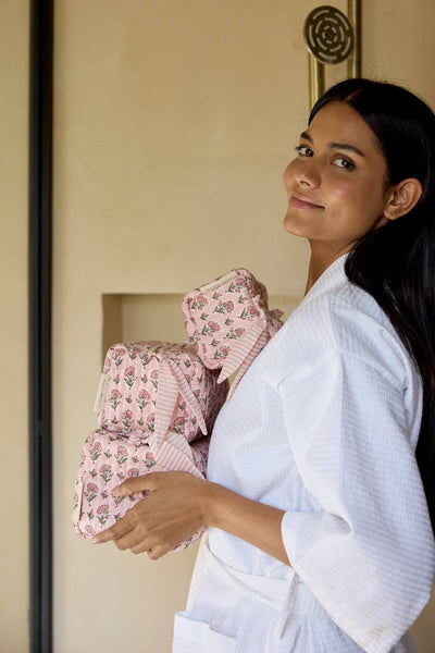 Woman in a white robe holding floral and striped cosmetic bags in a room.