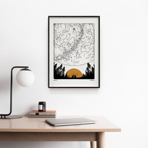 Framed artwork of a sunset over trees on a white wall above a desk.