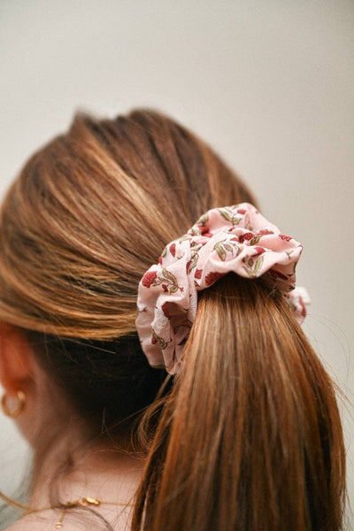 A close-up lifestyle shot of a woman with light brown hair wearing a dusty pink fabric scrunchie with a red and green floral pattern.