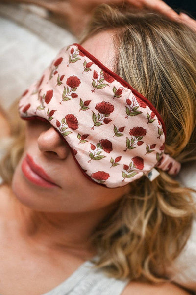 A close-up of a woman lying down wearing a light pink sleep mask with a red rose pattern and red trim.