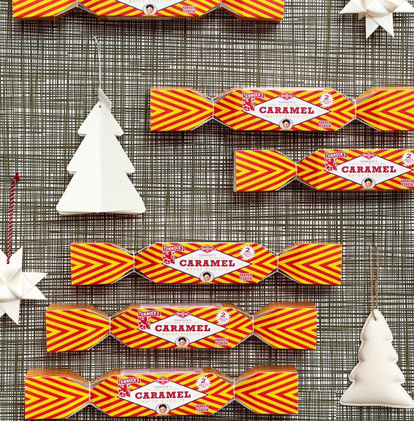 Five packages of Tunnock's brand Christmas crackers arranged on a grey textured surface, decorated with white paper ornaments.