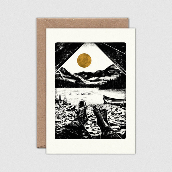 Greeting card with a black and white illustration of feet by a lake, a boat, and mountains, with a gold sun in the top right corner. The card is on a white background with a brown envelope.