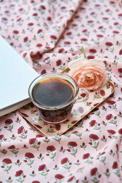 A styled flat-lay featuring a pink floral fabric with a repeating red rose and green leaf pattern. On the matching tray sits a clear glass cup filled with black coffee and a peach-pink rose.